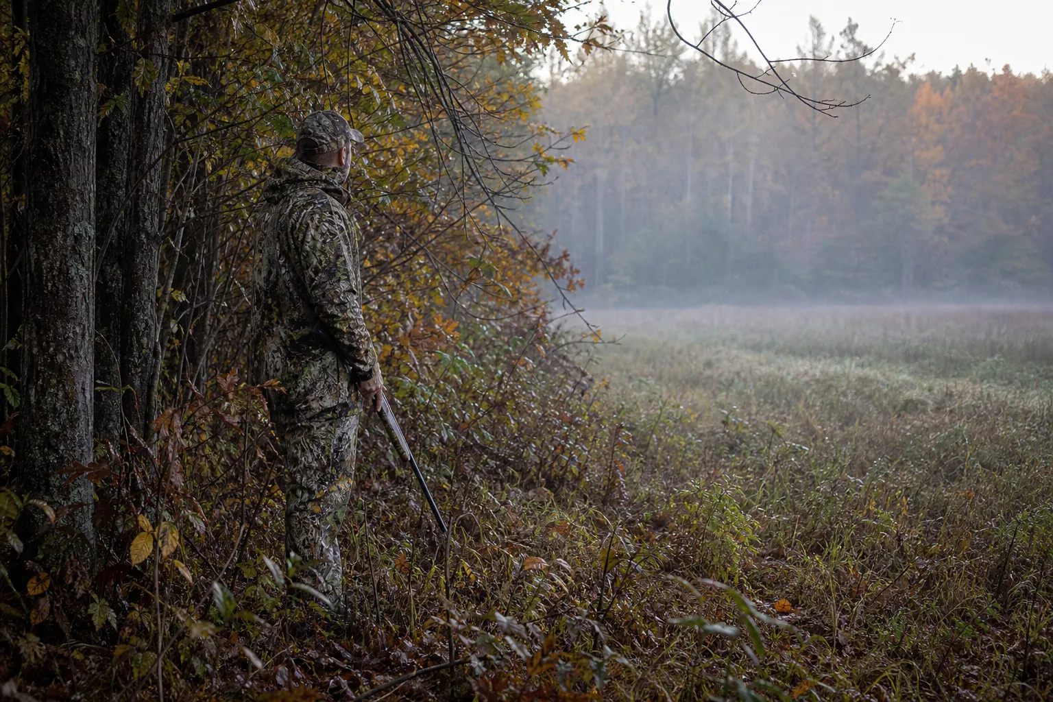 A camouflaged hunter stands at the edge of a wooded field at dawn with a rifle held safely pointed down, blending into the natural background.